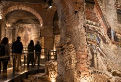 Visitors walk through a vaulted ancient Roman brick interior beside an exposed wall mosaic.